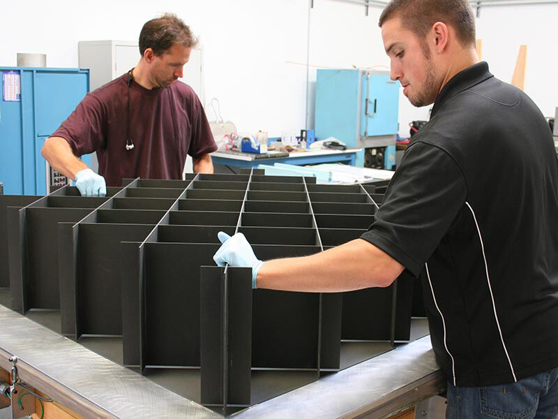 Image in a manufacturing environment. Two technicians are aligning a carbon fiber structure on top of a carbon fiber panel that is laying on a table with a steel surface.